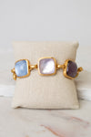 A gold-tone cuff bracelet with three large square cabochon glass stones in light blue, milky white, and dark purple, displayed on a beige fabric jewelry pillow over a white marble surface.