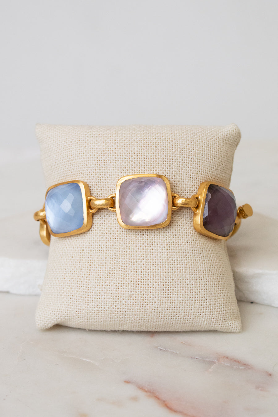 A gold-tone cuff bracelet with three large square cabochon glass stones in light blue, milky white, and dark purple, displayed on a beige fabric jewelry pillow over a white marble surface.