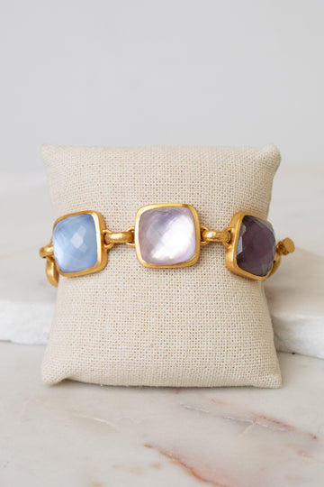 A gold-tone cuff bracelet with three large square cabochon glass stones in light blue, milky white, and dark purple, displayed on a beige fabric jewelry pillow over a white marble surface.
