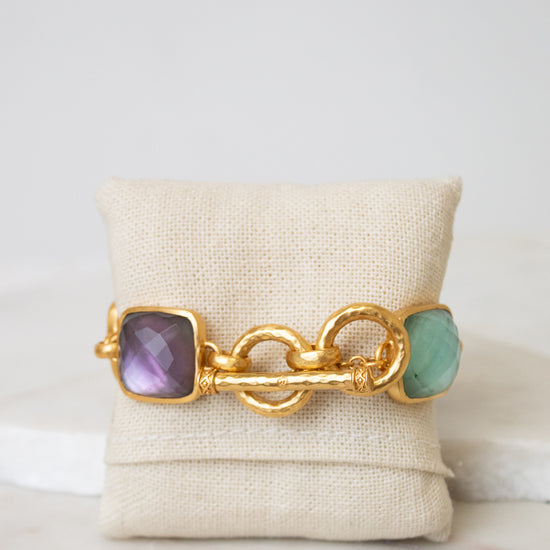 A gold chain-link bracelet shown from the toggle back closure showing multi-color square glass details on either end and displayed on a white canvas jewelry pillow
