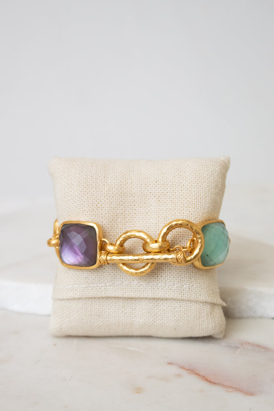 A gold chain-link bracelet shown from the toggle back closure showing multi-color square glass details on either end and displayed on a white canvas jewelry pillow