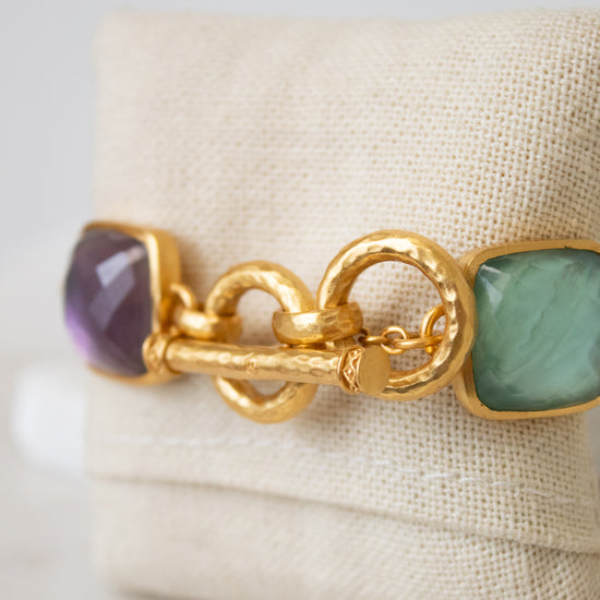 A gold-tone bracelet with a textured toggle clasp, featuring multi-color square glass details, displayed on a beige fabric jewelry stand.