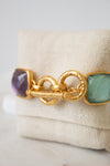 A gold-tone bracelet with a textured toggle clasp, featuring multi-color square glass details, displayed on a beige fabric jewelry stand.