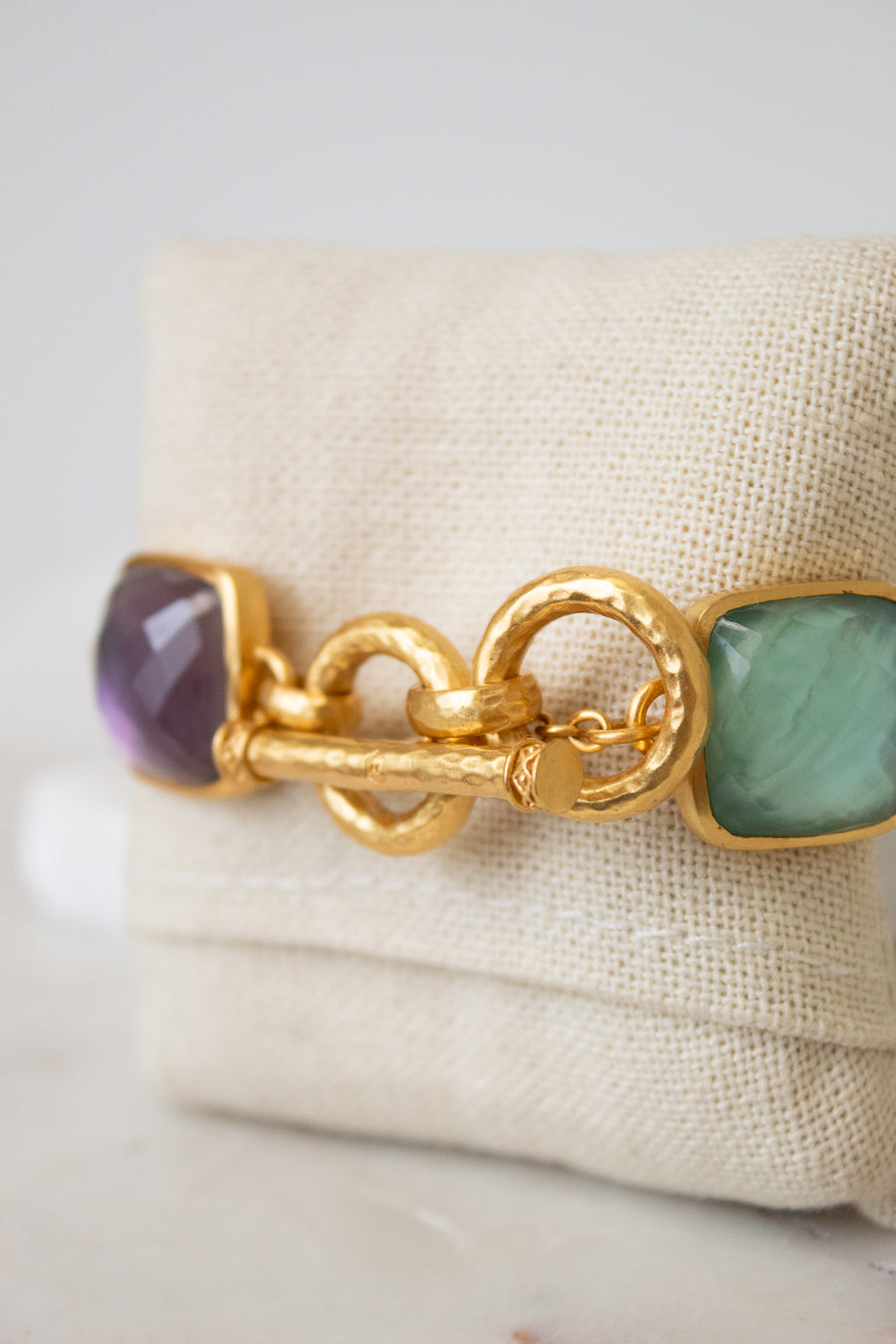 A gold-tone bracelet with a textured toggle clasp, featuring multi-color square glass details, displayed on a beige fabric jewelry stand.