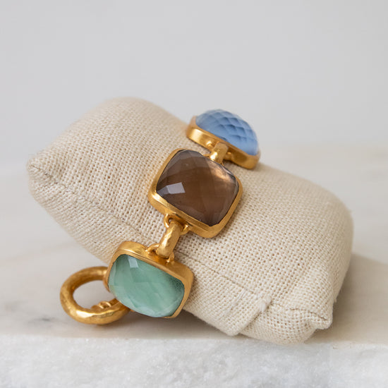 A gold-tone bracelet with multi-color square glass details resting on a beige fabric jewelry pillow over a white marble surface.