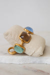 A gold-tone bracelet with multi-color square glass details resting on a beige fabric jewelry pillow over a white marble surface.