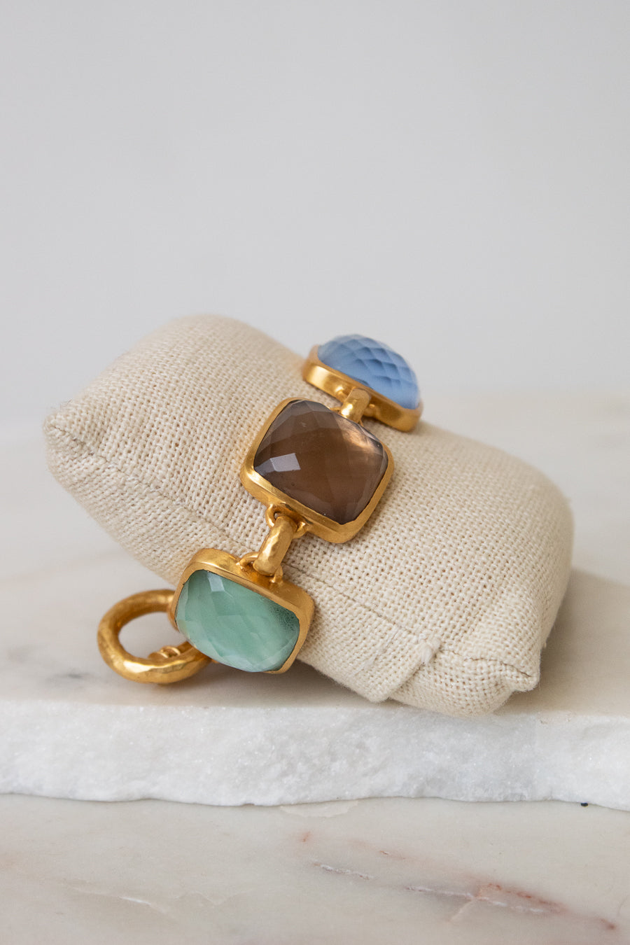 A gold-tone bracelet with multi-color square glass details resting on a beige fabric jewelry pillow over a white marble surface.