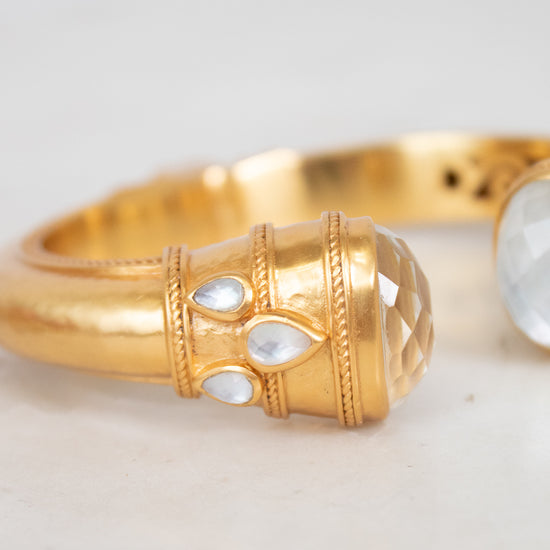 A close-up of a gold-toned ring featuring a textured cylindrical bezel set with three small clear teardrop-shaped stones and a larger faceted oval clear stone on the adjacent band, photographed on a light neutral surface.