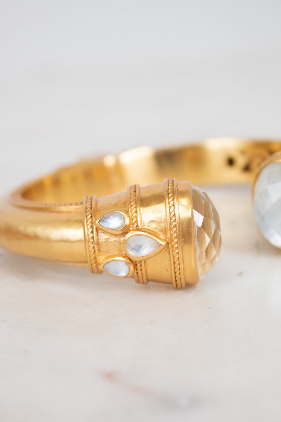 A close-up of a gold-toned ring featuring a textured cylindrical bezel set with three small clear teardrop-shaped stones and a larger faceted oval clear stone on the adjacent band, photographed on a light neutral surface.