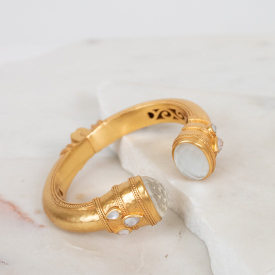 A gold-toned open cuff bracelet with carved interior details and white glass accents, displayed on a white marble surface against a light background.