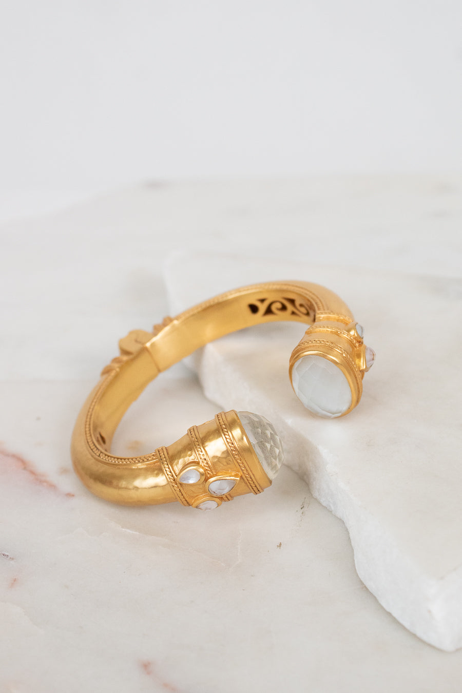 A gold-toned open cuff bracelet with carved interior details and white glass accents, displayed on a white marble surface against a light background.