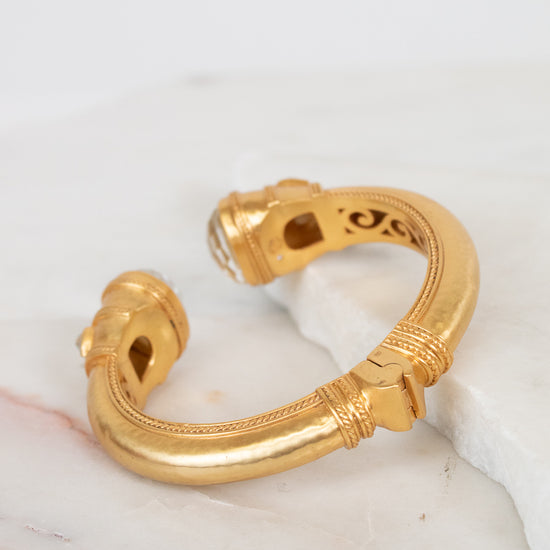 A gold-toned open cuff bracelet with decorative textured bands along outer edge and interior cutout details, displayed on a white marble surface.