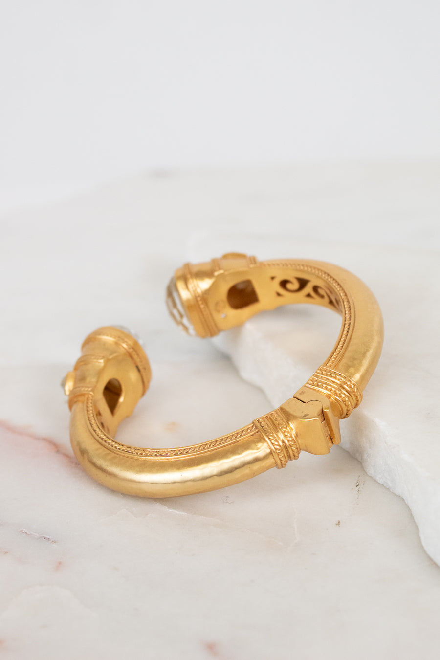 A gold-toned open cuff bracelet with decorative textured bands along outer edge and interior cutout details, displayed on a white marble surface.