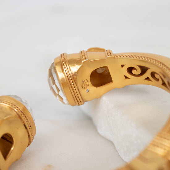 A close-up of a gold bangle bracelet with a white faceted glass stones set in gold-tone with an ornate interior filigree design, resting on a white marble block.