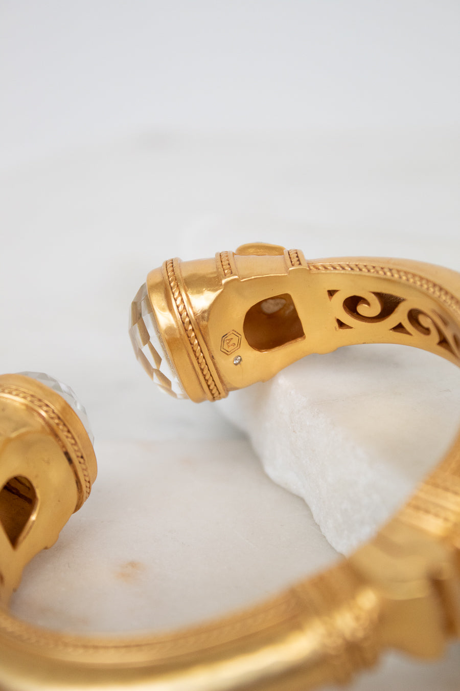 A close-up of a gold bangle bracelet with a white faceted glass stones set in gold-tone with an ornate interior filigree design, resting on a white marble block.