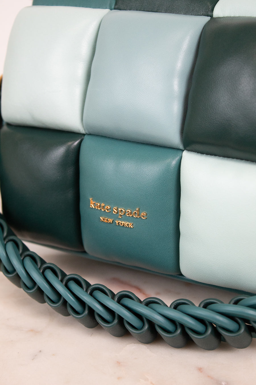 Close-up of a Kate Spade New York quilted leather handbag in shades of teal, dark forest green, and pale mint, featuring a braided shoulder strap and gold logo lettering.
