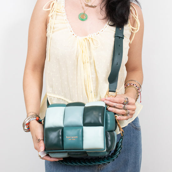 A woman wearing a cream tie-front blouse and blue jeans holds a green and white checkered leather shoulder bag with a braided strap. The brand name 'Kate Spade' is visible on the front of the bag.