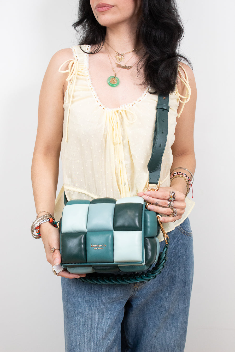 A woman wearing a cream tie-front blouse and blue jeans holds a green and white checkered leather shoulder bag with a braided strap. The brand name 'Kate Spade' is visible on the front of the bag.