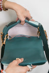 A teal leather shoulder bag with gold-tone hardware, shown open with a hand holding it open showing the interior. The bag features a dark teal interior of flap, a light colored fabric interior lining, and a dark teal shoulder strap.