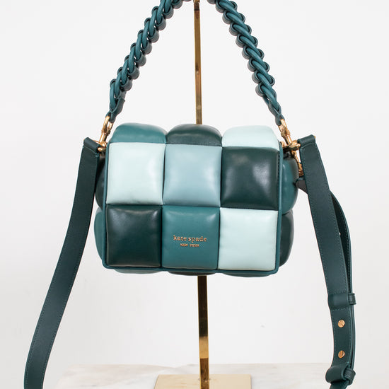 A woven leather shoulder bag by Kate Spade in shades of teal, mint green, and dark forest green, displayed on a gold stand with a marble surface against a white background.