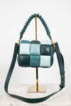 A woven leather shoulder bag by Kate Spade in shades of teal, mint green, and dark forest green, displayed on a gold stand with a marble surface against a white background.