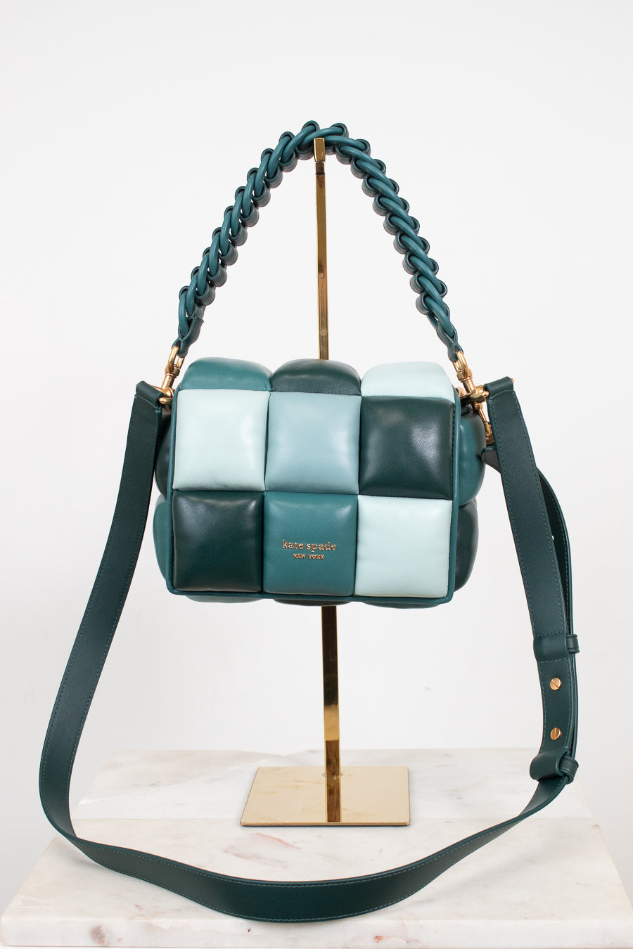 A woven leather shoulder bag by Kate Spade in shades of teal, mint green, and dark forest green, displayed on a gold stand with a marble surface against a white background.