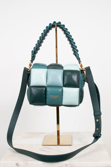 A woven leather shoulder bag by Kate Spade in shades of teal, mint green, and dark forest green, displayed on a gold stand with a marble surface against a white background.