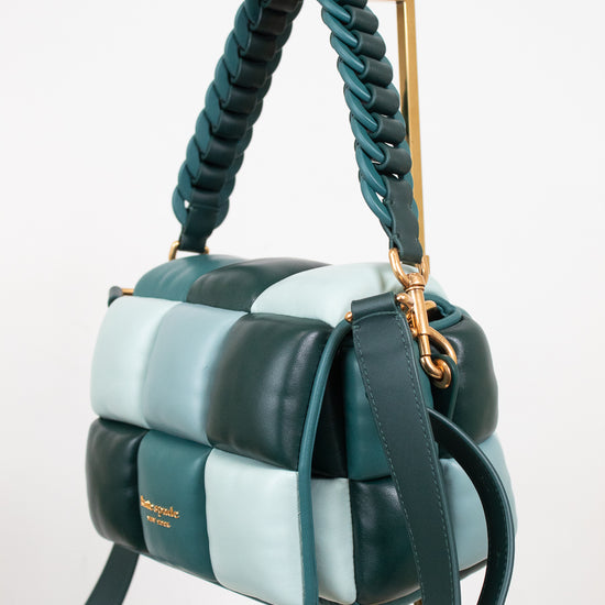 A structured handbag with a quilted block pattern in dark teal, light blue, and mint, featuring a braided top handle and a gold-tone stand on a white background.
