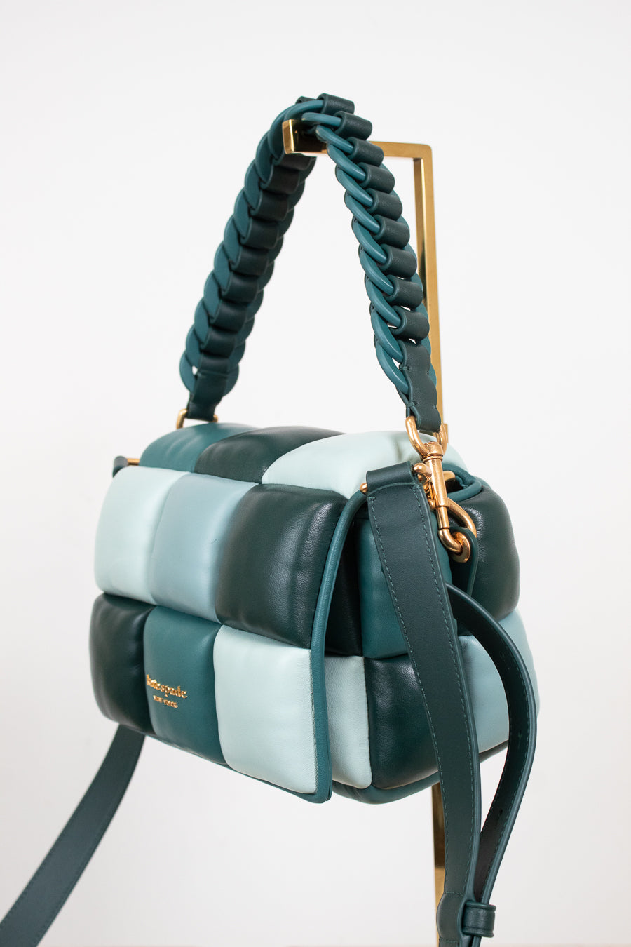 A structured handbag with a quilted block pattern in dark teal, light blue, and mint, featuring a braided top handle and a gold-tone stand on a white background.