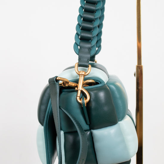 A structured handbag with a unique segmented, bubble-like design in shades of teal, dark green, and pale blue, featuring a thick braided top handle and gold-tone hardware, shown hanging against a white background on set on a gold stand.