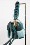 A structured handbag with a unique segmented, bubble-like design in shades of teal, dark green, and pale blue, featuring a thick braided top handle and gold-tone hardware, shown hanging against a white background on set on a gold stand.