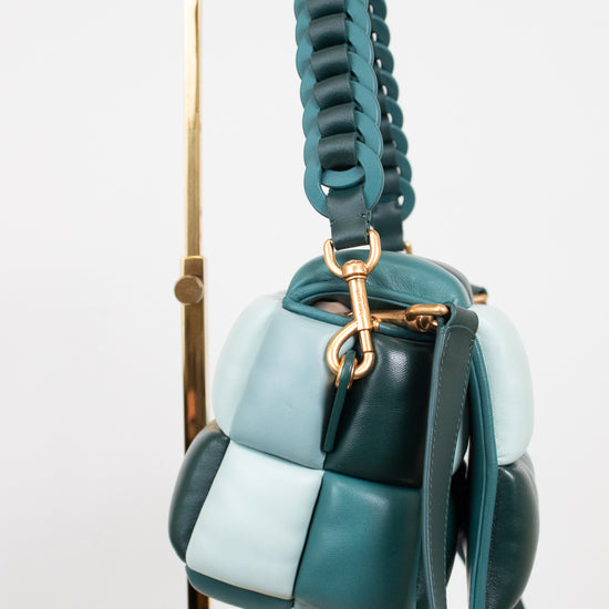 A small color-block leather handbag in shades of teal, dark green, and pale blue/mint, hanging from a gold metal display stand against a white background.