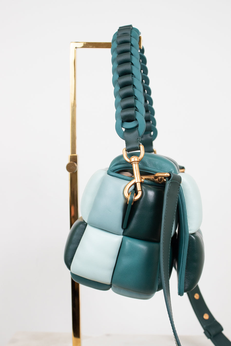 A small color-block leather handbag in shades of teal, dark green, and pale blue/mint, hanging from a gold metal display stand against a white background.