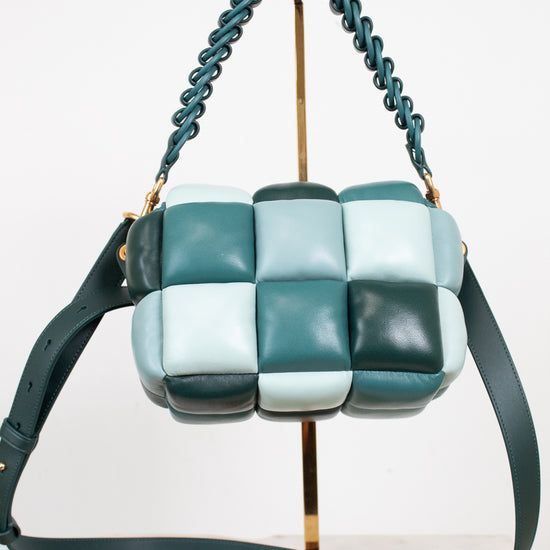 A small color-block leather shoulder bag with a woven checkerboard pattern in shades of teal, mint, and green, featuring a teal braided strap and a matching shoulder strap, displayed on a gold stand.