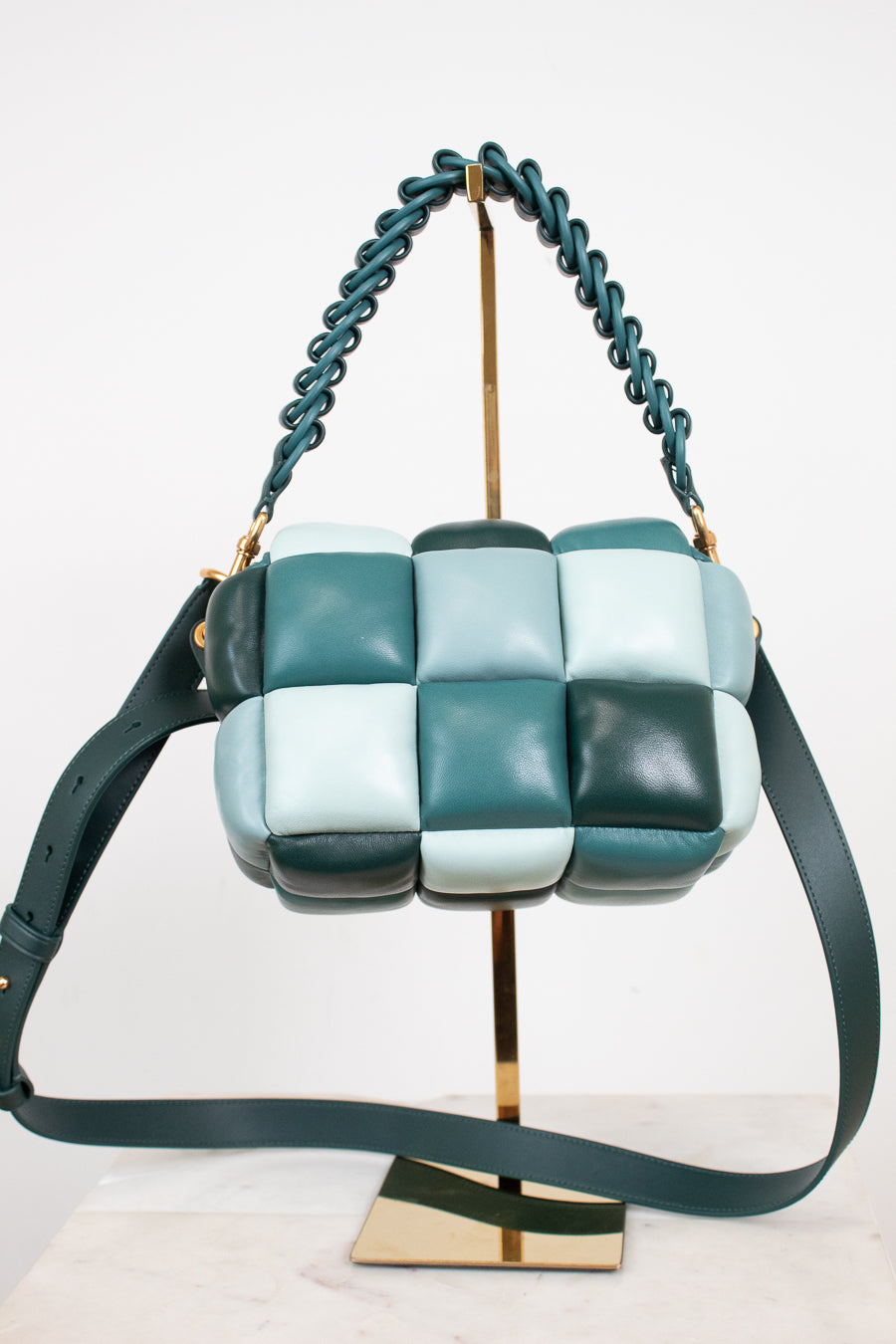 A small color-block leather shoulder bag with a woven checkerboard pattern in shades of teal, mint, and green, featuring a teal braided strap and a matching shoulder strap, displayed on a gold stand.