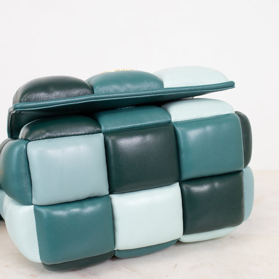 A modern cube-shaped color-block handbag with a padded, segmented leather design in a checkered color-block pattern of teal, dark forest green, and pale mint, resting on a light marble surface against a white background.