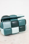 A modern cube-shaped color-block handbag with a padded, segmented leather design in a checkered color-block pattern of teal, dark forest green, and pale mint, resting on a light marble surface against a white background.