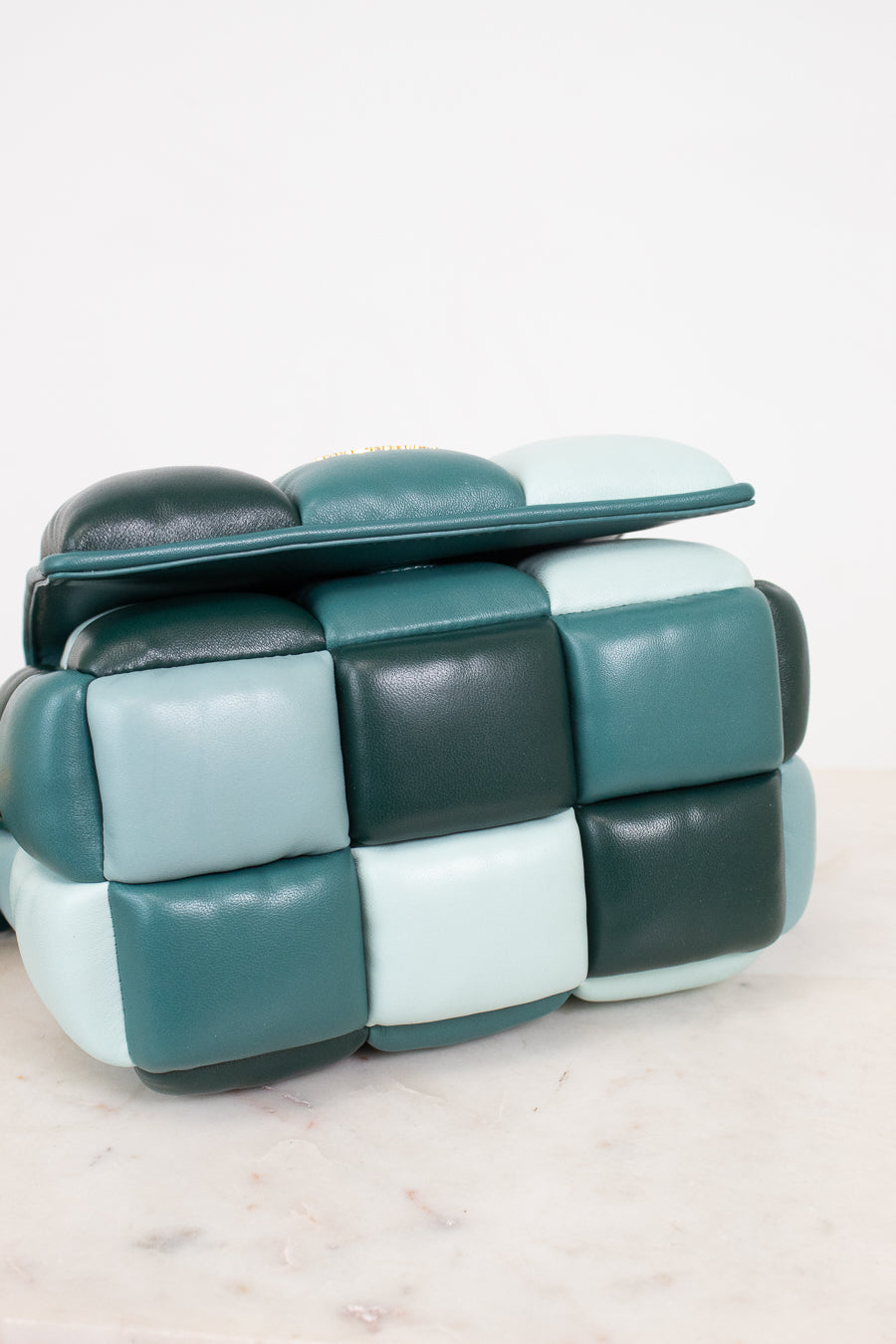 A modern cube-shaped color-block handbag with a padded, segmented leather design in a checkered color-block pattern of teal, dark forest green, and pale mint, resting on a light marble surface against a white background.