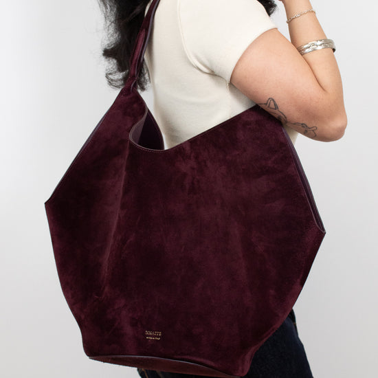 A woman wearing a cream t-shirt and denim shorts carries a large, deep burgundy suede shoulder bag against a plain light gray background.