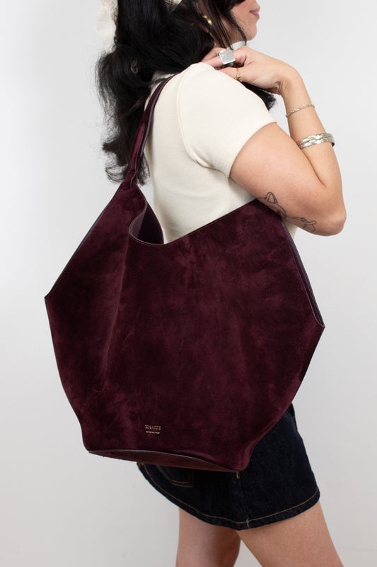 A woman wearing a cream t-shirt and denim shorts carries a large, deep burgundy suede shoulder bag against a plain light gray background.