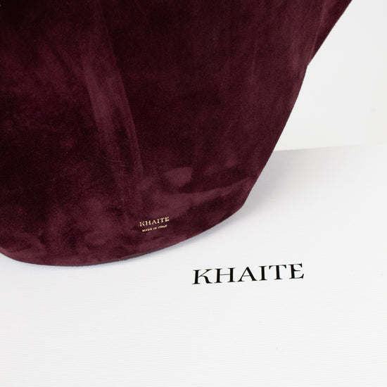 A close-up of a suede shoulder bag in deep burgundy, resting on top of a white box with the brand name 'KHAITE' printed in black.