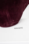 A close-up of a suede shoulder bag in deep burgundy, resting on top of a white box with the brand name 'KHAITE' printed in black.
