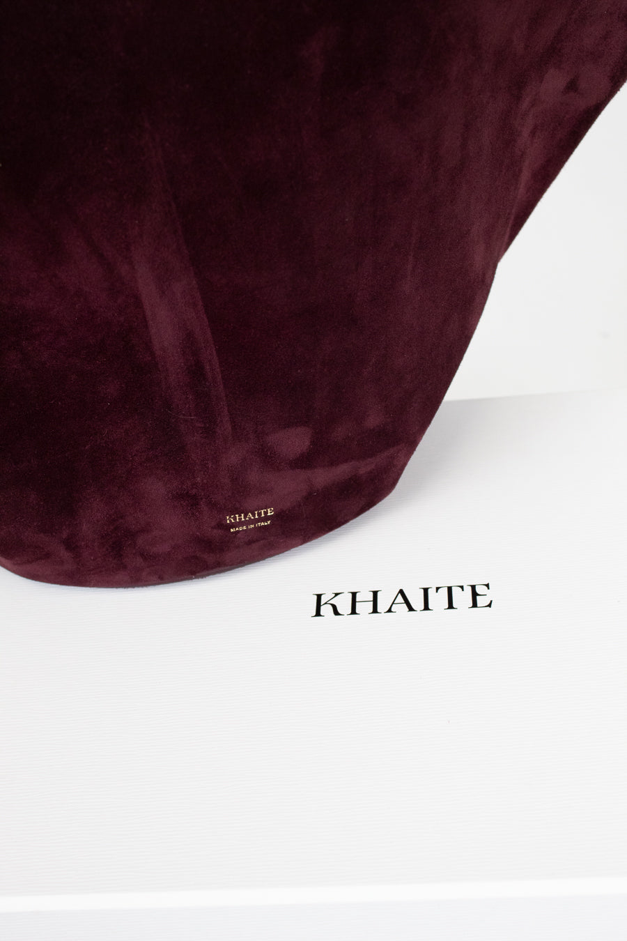 A close-up of a suede shoulder bag in deep burgundy, resting on top of a white box with the brand name 'KHAITE' printed in black.