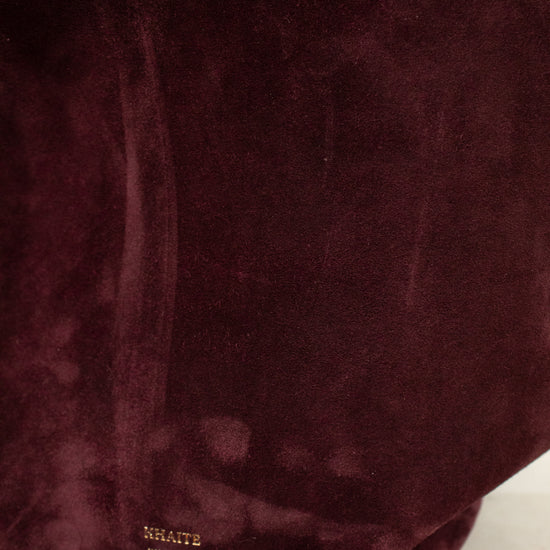 A close-up of a deep burgundy suede fabric surface with a small gold 'KHAITE' logo near the bottom, resting on a light marble surface.