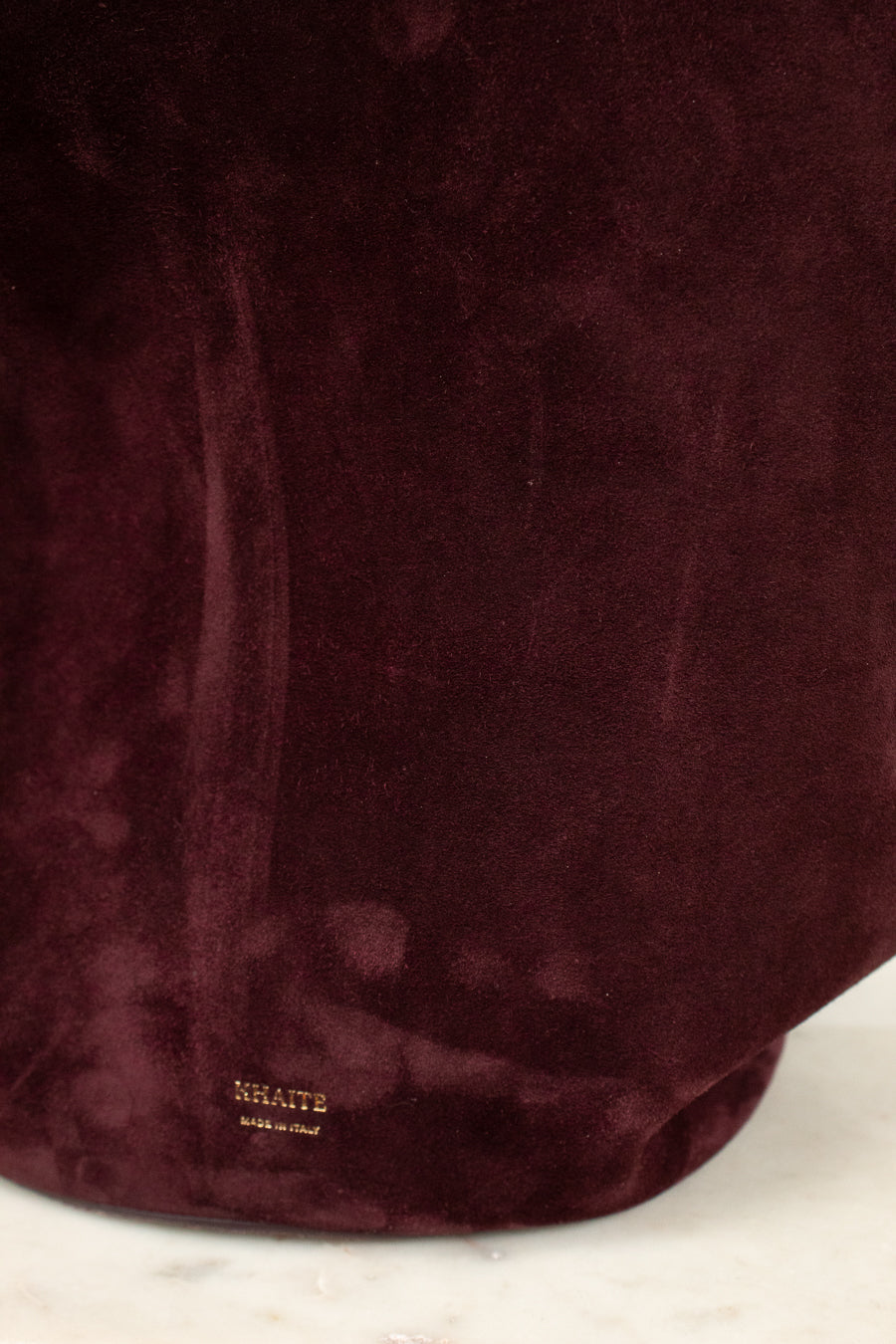 A close-up of a deep burgundy suede fabric surface with a small gold 'KHAITE' logo near the bottom, resting on a light marble surface.