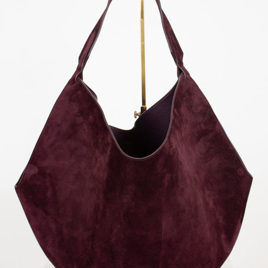 A large slouchy hobo shoulder bag in deep burgundy suede, displayed on a white surface against a white background with a gold stand visible behind it.