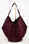 A large slouchy hobo shoulder bag in deep burgundy suede, displayed on a white surface against a white background with a gold stand visible behind it.