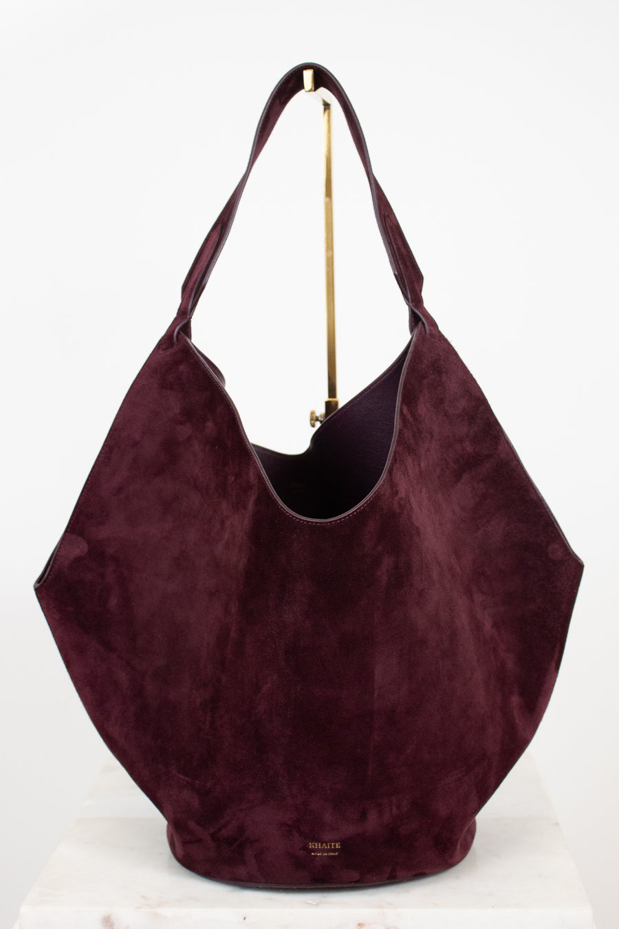 A large slouchy hobo shoulder bag in deep burgundy suede, displayed on a white surface against a white background with a gold stand visible behind it.