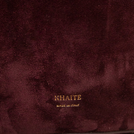 Close-up of a deep burgundy suede fabric surface with a small gold KHAITE logo and 'MADE IN ITALY' text near the bottom center.