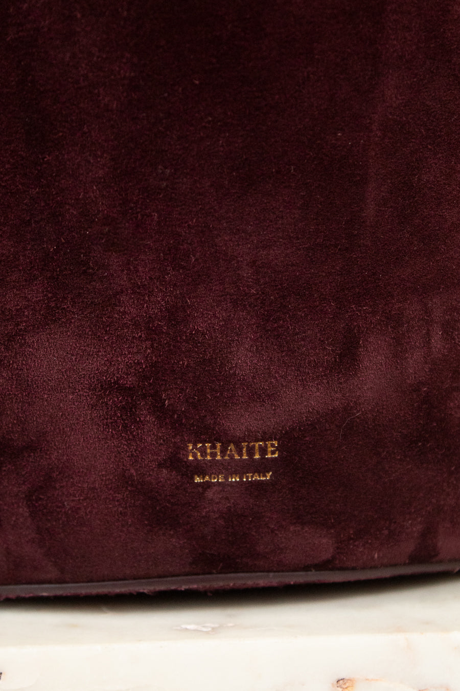 Close-up of a deep burgundy suede fabric surface with a small gold KHAITE logo and 'MADE IN ITALY' text near the bottom center.